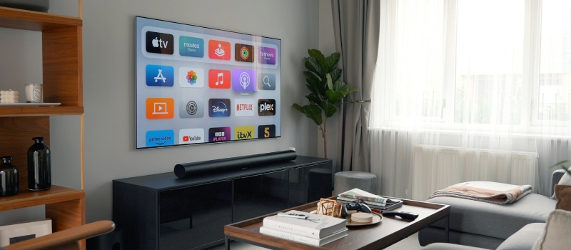 Wykorzystaj w pelni mozliwosci swojego telewizora dzieki soundbarowi firmy Vebos. telewizor na scianie, pod ktorym znajduje sie soundbar na czarnym meblu pod telewizor, po lewej stronie telewizora drewniana szafka, a po prawej zielona roslina